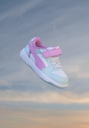 Baskets enfant blanches et roses avec bride en velcro et lacets flottant sur un fond de ciel bleu doux et beige.