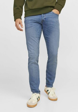 Man wearing light blue slim-fit jeans, white sneakers with green stripes, and olive green sweater with one hand in pocket.