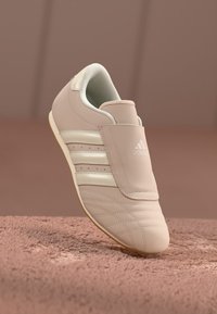 Lys beige Adidas-sneaker med tre hvite striper og et dekket snøringsområde, plassert på en strukturert brun overflate.