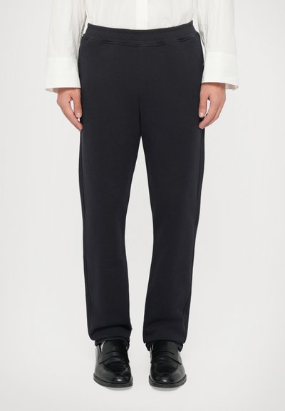 Personne portant un pantalon noir ajusté avec des chaussures de ville noires et une chemise blanche à manches longues, se tenant devant un fond uni.