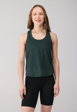 Mladá žena sa usmieva, má na sebe tmavozelený tielko bez rukávov a čierne priliehavé krátke nohavice, stojí pred jednofarebným svetlosivým pozadím.