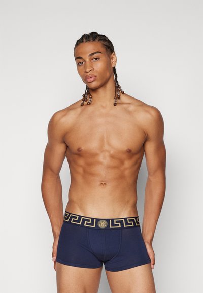 Marineblå herreboxershorts med en sort og gul logoelastik med et græsk nøglemønster. Stoffet har en glat tekstur.