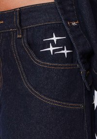 Vaqueros de denim oscuro con costuras naranjas, un bolsillo delantero y acentos bordados en blanco que parecen estrellas en el bolsillo trasero.