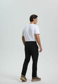 Mies nähtävissä takaa päällä valkoinen t-paita, mustat cargo-housut ja vaaleat vaelluskengät seisomassa vaaleaa taustaa vasten.