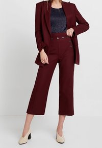 Weinroter, maßgeschneiderter Blazer mit einem Knopf, hoch taillierte, kurze Hose mit Doppelknöpfen, getragen über einem dunklen, funkelnden Oberteil und beige Absätze.