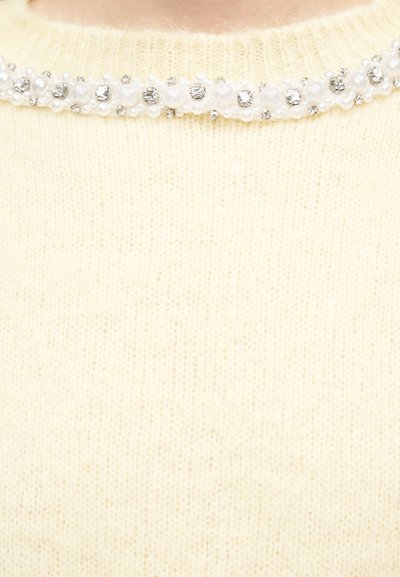 Λαιμόκοψη πλεκτού πουλόβερ σε κρεμ χρώμα, διακοσμημένη με σειρά εναλλασσόμενων μαργαριταριών και μικρών διαφανών στρας.