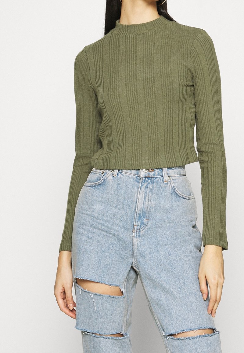 Top de punto acanalado en verde oliva con cuello alto, mangas largas y un ajuste corto, combinado con jeans desgastados de color azul claro.
