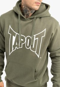 Olīvu zaļš kapucju džemperis, kas izgatavots no kokvilnas un sintētiskā auduma, ar baltu "TAPOUT" logotipu, auklītēm un priekšējo kangaru kabatu.