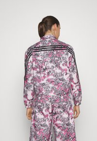 Tuta sportivo in un vivace rosa, nero e bianco con un motivo astratto. Presenta polsini elasticizzati, un colletto e tre strisce nere sul retro.