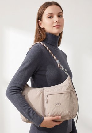 Jonge vrouw in een grijze coltrui houdt een beige schoudertas met het GMV-logo en een patroonriem vast tegen een effen achtergrond.