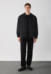 Chaqueta ligera negra con dos bolsillos en el pecho, combinada con una camiseta y pantalones negros. El modelo lleva zapatos negros gruesos; diseño minimalista.