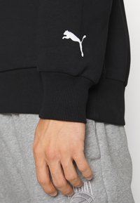 Ręka oparta w pobliżu kieszeni szarych dresów, ubrana w czarny sweatshirt z podwiniętymi rękawami i białym logo Puma na nadgarstku.