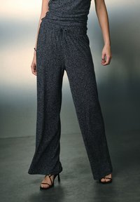 Zwarte, glinsterende broek met wijde pijpen en een trekkoord in de taille. Ze hebben een gladde textuur en een losse pasvorm, gecombineerd met zwarte sandalen met bandjes.