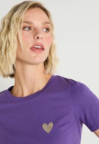 Camiseta morada con cuello redondo y mangas cortas. Presenta un pequeño aplique de corazón dorado en el lado izquierdo del pecho. Textura de tela suave.