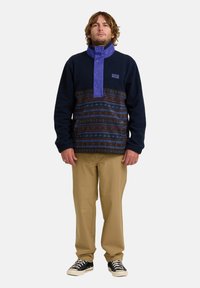 Fleece pullover in marineblauw met paarse kraag en knopen, met een gemusterde middensectie in blauwe en bruine strepen. Gedragen met beige broek.