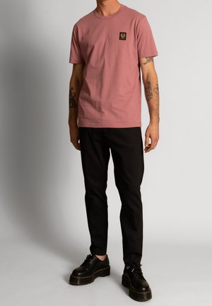 Lyserød bomuld t-shirt med rund halsudskæring og sort logo-patch, kombineret med sorte bukser og sorte snørelace-sko.