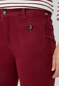 Nahaufnahme einer Person, die dunkelrote Jeans mit einer Knopfleiste und Taschenverzierungen trägt, kombiniert mit einem weiß-rot gestreiften Oberteil.