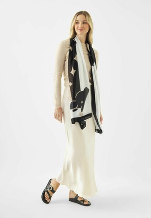 Woman wearing beige sweater, long cream skirt, black and white patterned scarf, and black sandals, standing against a white background.