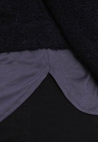 Marinblått fluffigt tröja över en grå skjorta med rundad nederkant, med kontrasterande texturer och mörk tyg under, vilket ger intryck av en lagerlook.