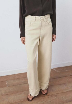 Persoon die beige spijkerbroeken met hoge taille en wijde pijpen met omgeslagen zomen draagt, een donker lange mouwen top en bruine slippers, binnen staand.