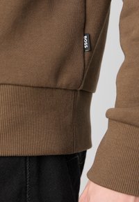 Sweatshirt marron avec ourlet et poignets côtelés, avec une petite étiquette noire « BOSS », porté par-dessus un pantalon noir sur le côté d’une personne.