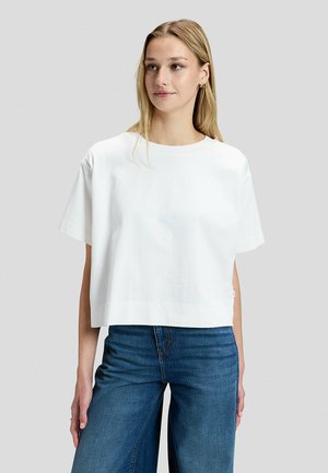 Młoda kobieta ubrana w luźny biały t-shirt z krótkim rękawem i niebieskie szerokie dżinsy, stoi i patrzy lekko na bok na jednolitym tle.