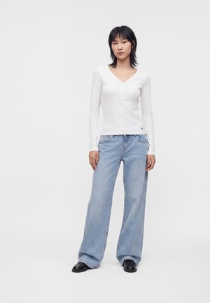 Persona in piedi di fronte, indossa una maglia bianca aderente a maniche lunghe con scollo a V, jeans azzurri a gamba larga e scarpe nere su sfondo semplice.