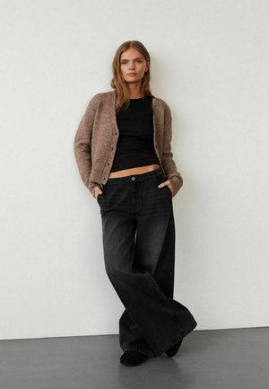 Brun cardigan over en sort top, parret med vide ben sorte jeans. Cardiganen har knapper, og jeansene har høj talje og løs pasform.