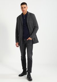 Mørk grå strikket frakk med sjalskrage og knappelukking, kombinert med en marineblå polo, mørke jeans og sorte ankelstøvler.