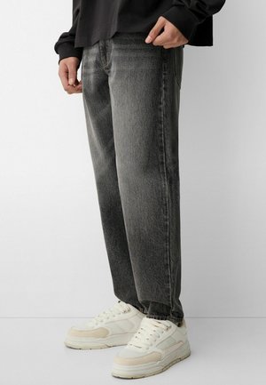 Sötétszürke, laza szabású farmernadrág, denim anyagból készült. Diszkrét kopásokkal és kontrasztos varrással rendelkezik. Fehér és krém színű sportcipővel párosítva.
