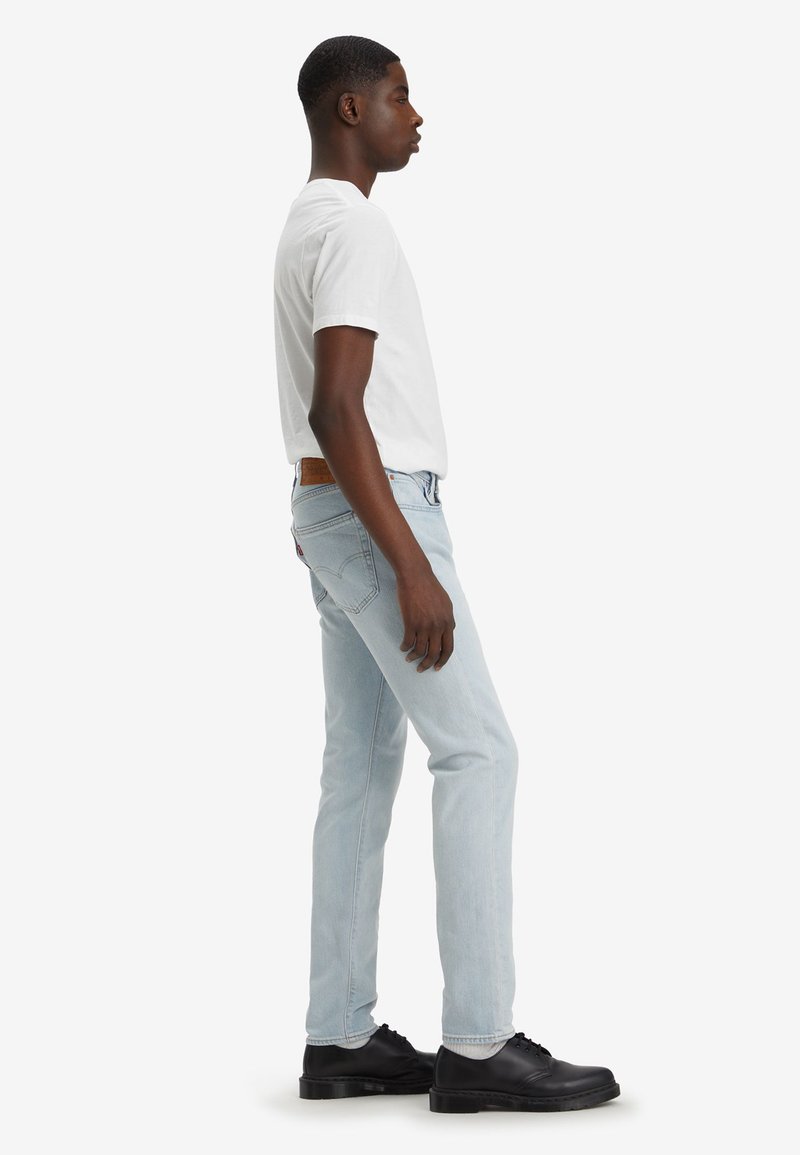 Jean de mezclilla azul claro con un ajuste slim, material de algodón, diseño de cinco bolsillos, parche de cuero marrón en la cintura trasera, combinados con zapatos negros.