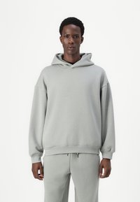 Lys grå hettegenser laget av myk stoff; har en stor hette, oversized passform, senkede skuldre og ribbestrikkede mansjetter. Kombinert med matchende joggebukser.