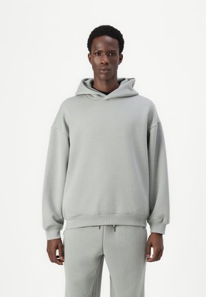 Felpa grigia chiaro realizzata in un tessuto morbido; presenta un ampio cappuccio, vestibilità oversize, spalle scese e polsini a costine. Abbinata a pantaloni della tuta coordinati.