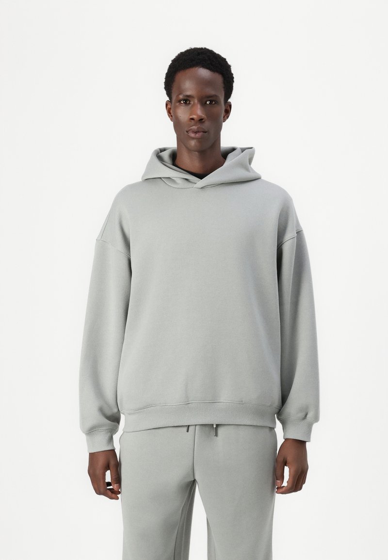 Lys grå hettegenser laget av myk stoff; har en stor hette, oversized passform, senkede skuldre og ribbestrikkede mansjetter. Kombinert med matchende joggebukser.