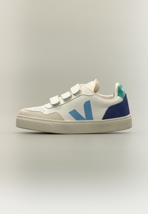 Witte sneaker met blauw "V"-logo, drie klittenbandsluitingen, beige suède details en donkerblauwe en groene hielaccenten op een effen achtergrond.