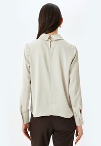 Blusa de un beige claro con mangas largas, cuello y cierre de botones en la parte trasera. Presenta una textura suave y lisa con un ligero brillo.