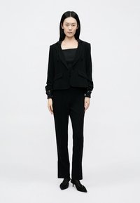 Must ülikond, millel on kapuutsiga lühike jakk, kaks esitaskut ja vormitud püksid, kõik siledast kangast ning kitsas lõikega.