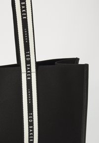 Schwarze Tragetasche aus strukturiertem Stoff mit einem weißen und schwarzen gestreiften Tragegurt und geprägt Logos von "Ted Baker London".