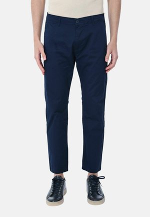 Chino in cotone blu navy con vestibilità slim, dotati di chiusura a bottone, passanti per cintura e tasche laterali. Indossati con sneakers nere.
