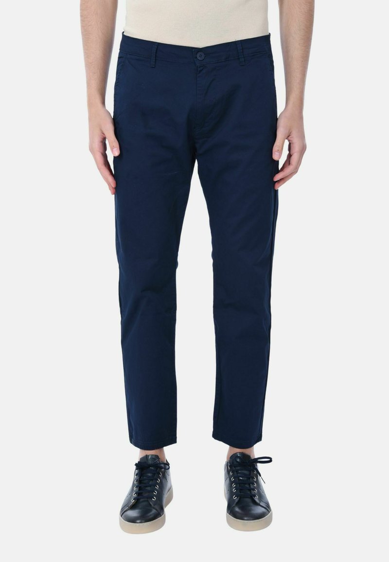 Chino in cotone blu navy con vestibilità slim, dotati di chiusura a bottone, passanti per cintura e tasche laterali. Indossati con sneakers nere.