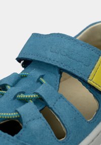 Μπλε σανδάλι από τσόχα με κίτρινα στοιχεία, ανοιχτές πλευρές και ραμμένες λεπτομέρειες. Διαθέτει ιμάντα με βέλκρο και στρογγυλεμένη λευκή σόλα για άνεση.
