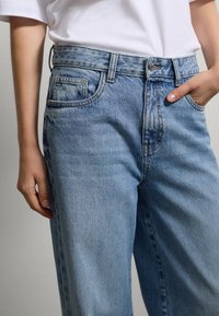Persona che indossa jeans di denim azzurro chiaro e una camicia bianca, con una mano in tasca e l'altra rilassata al suo fianco.