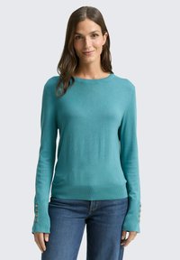 Türkisfarbener Strickpullover mit langen Ärmeln, Rippbündchen mit goldfarbenen Knopfakzenten und rundem Halsausschnitt. Kombiniert mit blauen Jeans.