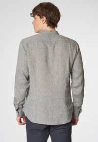 Un giovane visto di spalle, che indossa una camicia di lino grigia chiara con maniche lunghe, in piedi contro uno sfondo bianco semplice.