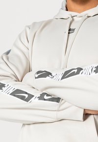 Ljus beige hoodie med en grå Nike-logotyp på bröstet och tryckta ärmdekorationer. Tyget verkar mjukt med en slät yta.