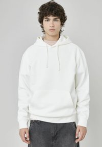 Sweatshirt à capuche blanche avec une poche avant, des cordons de serrage et des manches longues, porté par-dessus une chemise rose clair, sur un fond neutre.