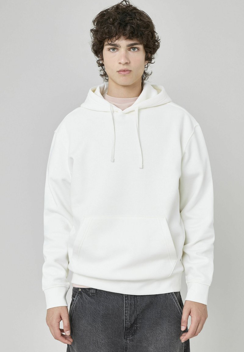 Sweatshirt à capuche blanche avec une poche avant, des cordons de serrage et des manches longues, porté par-dessus une chemise rose clair, sur un fond neutre.