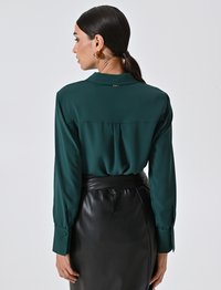 Donna con capelli scuri raccolti in una coda di cavallo bassa, che indossa orecchini a cerchio dorati, una blusa a maniche lunghe verde scuro e una gonna di pelle nera a vita alta.