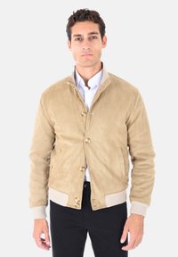 Uomo che indossa una giacca beige scamosciata con bottoni sopra una camicia bianca e pantaloni neri, in piedi contro uno sfondo chiaro uniforme.