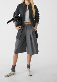 Grauer Reißverschluss-Pullover, graue weit geschnittene, gefaltete Shorts, silberne Sneaker und schwarze Handtasche. Schlichtes Design mit glatter Textur.